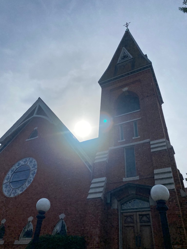 The sun setting behind a brick church with a cross ascending toward the sky.