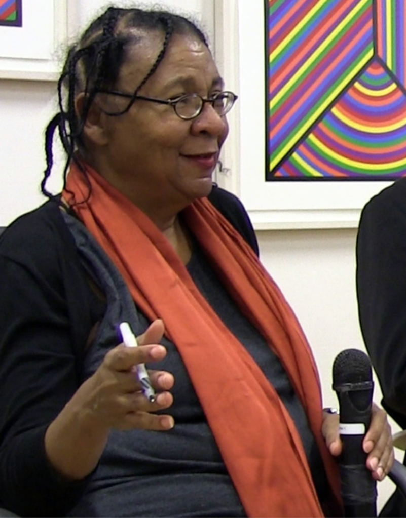 bell hooks seated on a panel with a microphone in one hand and a pen in another.